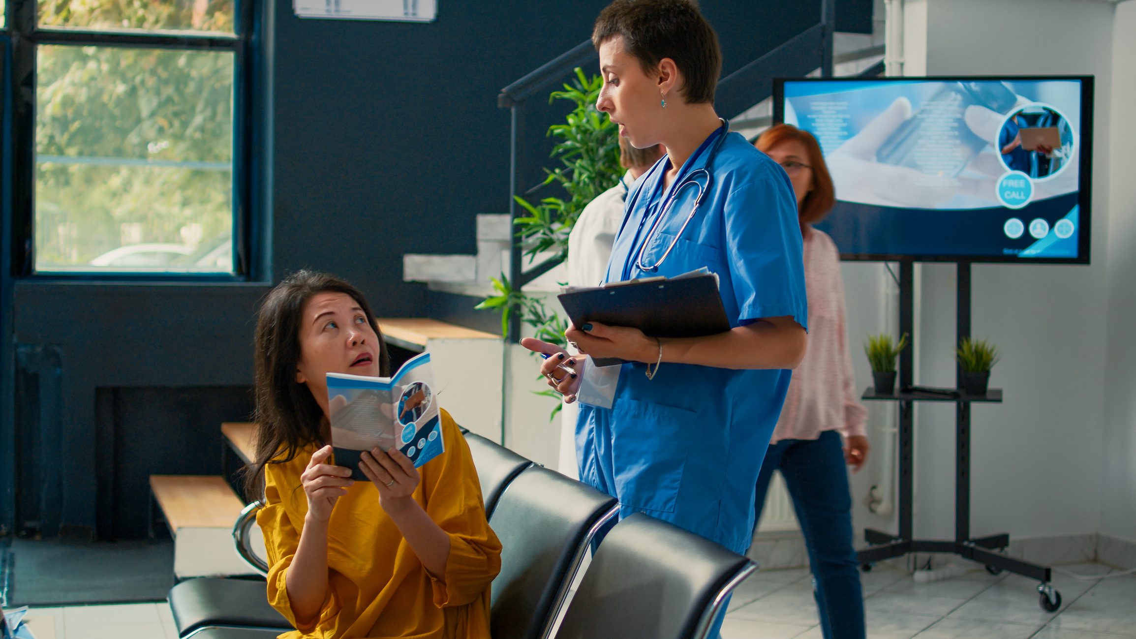 Medical assistant talking to asian patient in hospital lobby, looking at medical form to receive healthcare insurance at checkup visit. Nurse consulting elderly woman in waiting area.