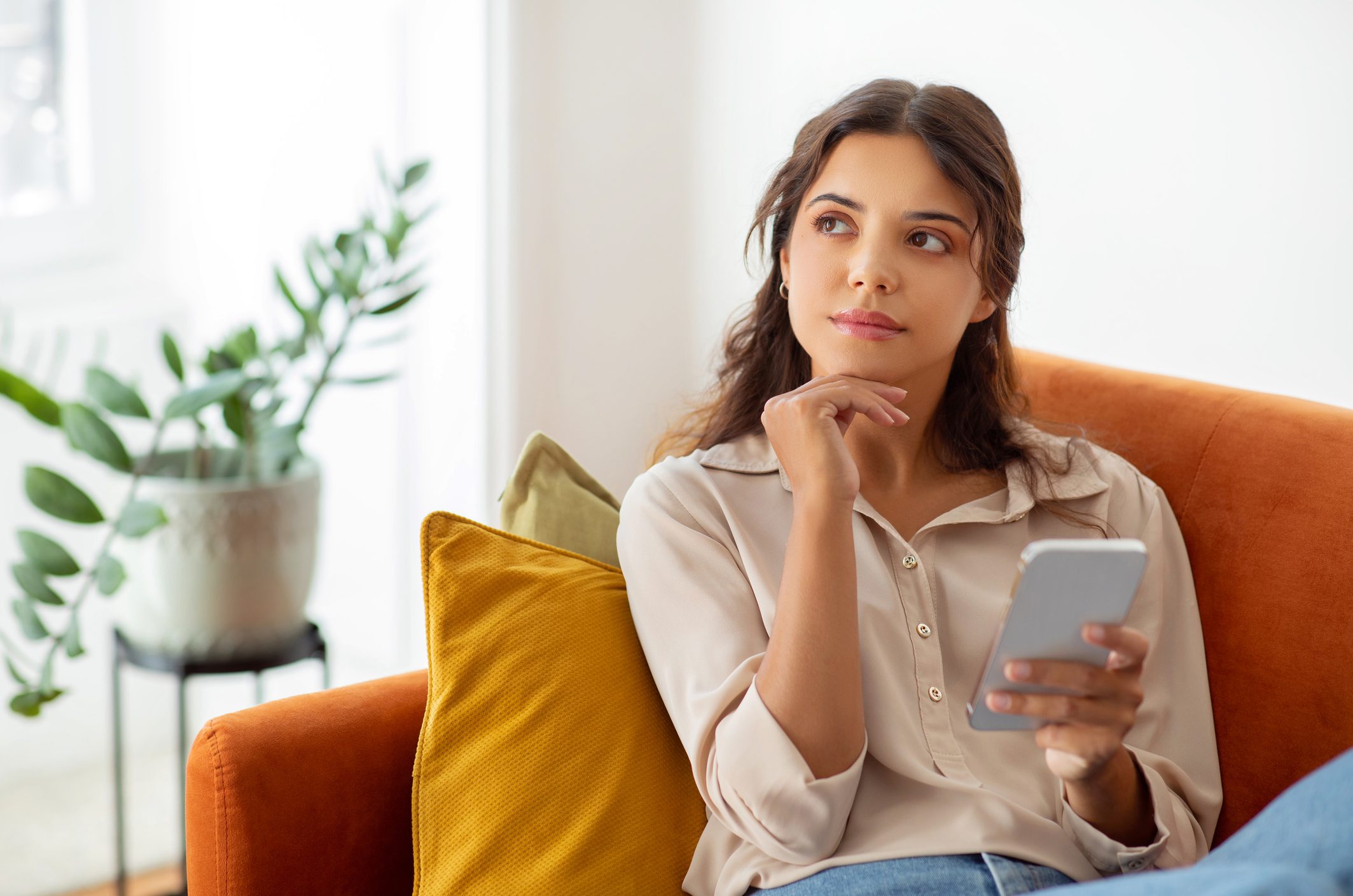 Pensive young woman holding smartphone and looking away while relaxing on couch at home, beautiful female deep in thoughts using mobile phone in domestic interior, thinking and planning something