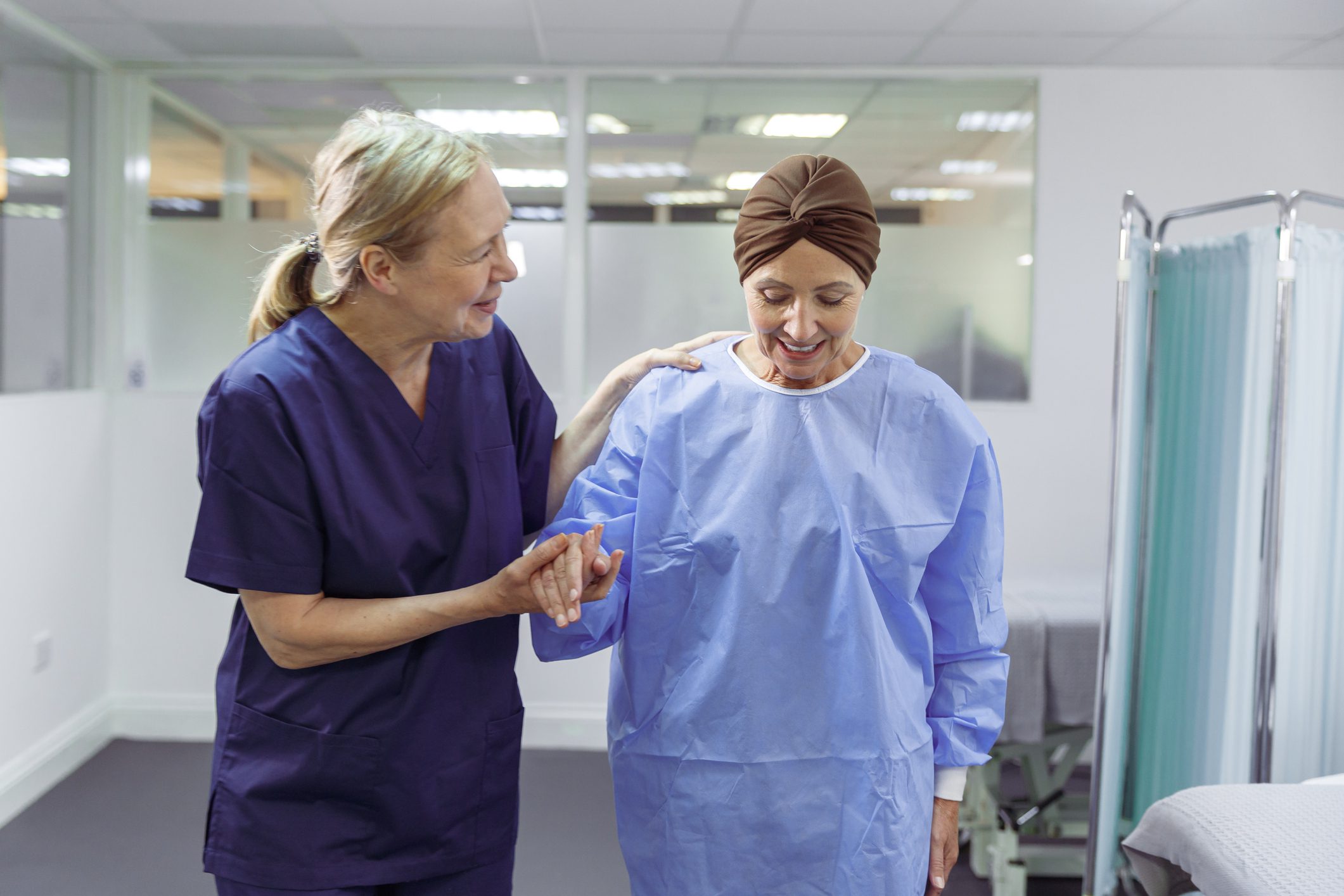A caring mature adult nurse holds hands with and helps support a female patient as she walks through the hospital ward. The patient is a woman with cancer who is undergoing chemotherapy treatment.