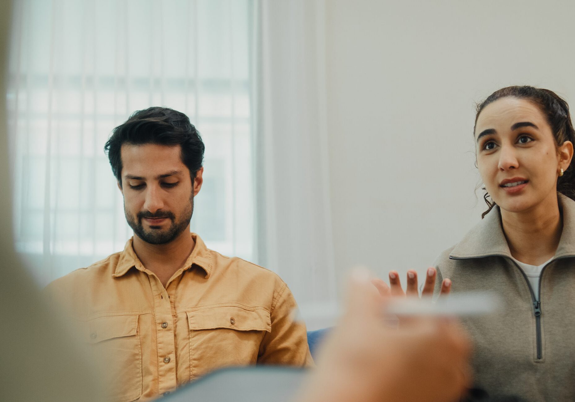 Closeup of young couple in serious discussion with mental health professional during marriage counseling session sit on sofa in living room at home. Therapy session and mental health concept.