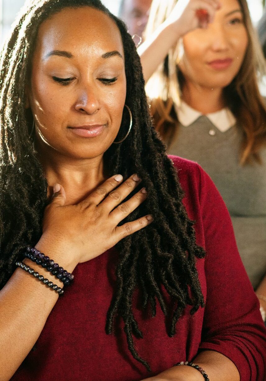 A diverse group of people in a workshop setting, focusing on mindfulness. Diverse are seated, with one woman in the foreground practicing a calming exercise with hand on her chest.